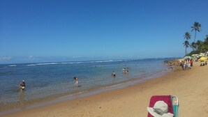 Una spiaggia con persone che nuotano e passeggiano sulla riva, palme e ombrelloni.