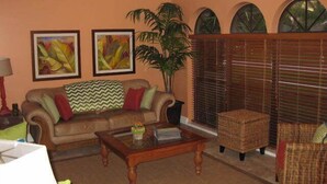 Una sala de estar con un sofá marrón, una mesa de centro de madera y una silla de mimbre. Hay dos cuadros en la pared y una planta grande.