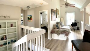 A spacious room with a white staircase, a bed with multiple pillows, a desk with a chair, and a bookshelf.