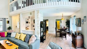 A spacious living area with a grey sofa, a wooden coffee table, and a dining table with chairs. There is a staircase leading to an upper level.