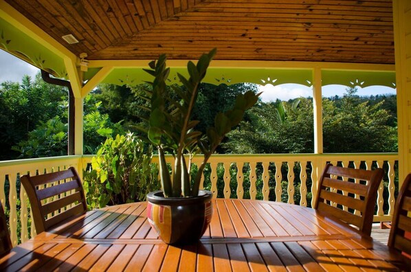 A covered outdoor seating area with a wooden table and chairs, surrounded by lush greenery.