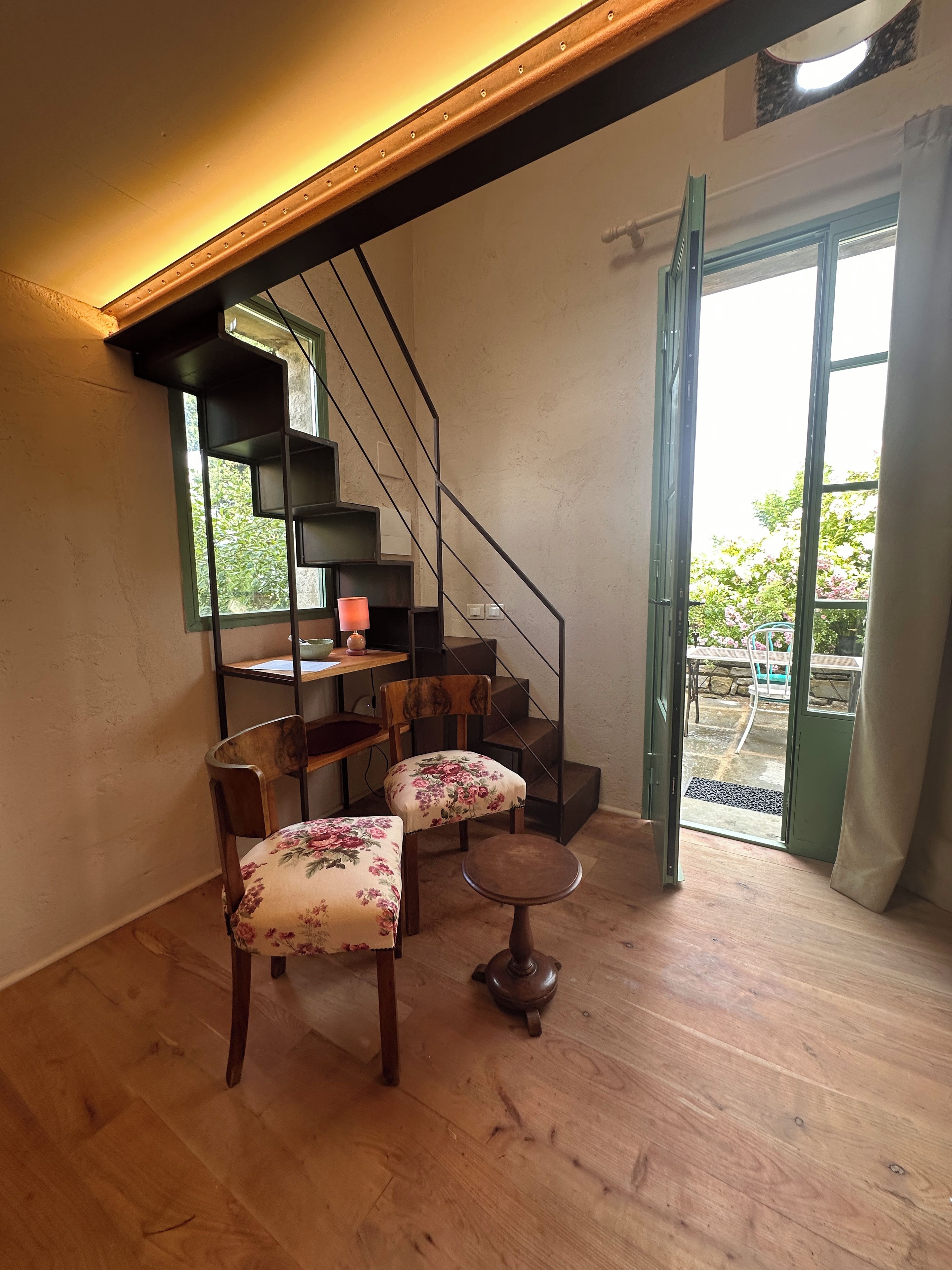 A room with a wooden floor, a staircase, a desk with a lamp, two chairs with floral upholstery, and a view through a glass door.