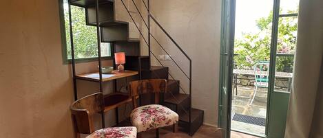A room with a wooden floor, a staircase, a desk with a lamp, two chairs with floral upholstery, and a view through a glass door.