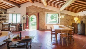 A spacious dining area with a wooden table and chairs, a kitchen in the background, and a view of the outdoors through a large window.