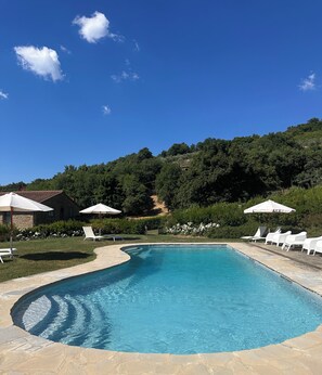 A swimming pool with white lounge chairs and umbrellas, surrounded by greenery and a stone building.