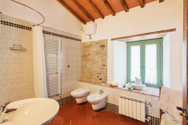A bathroom with a bathtub, two toilets, a bidet, a window with shutters, and a stone wall.