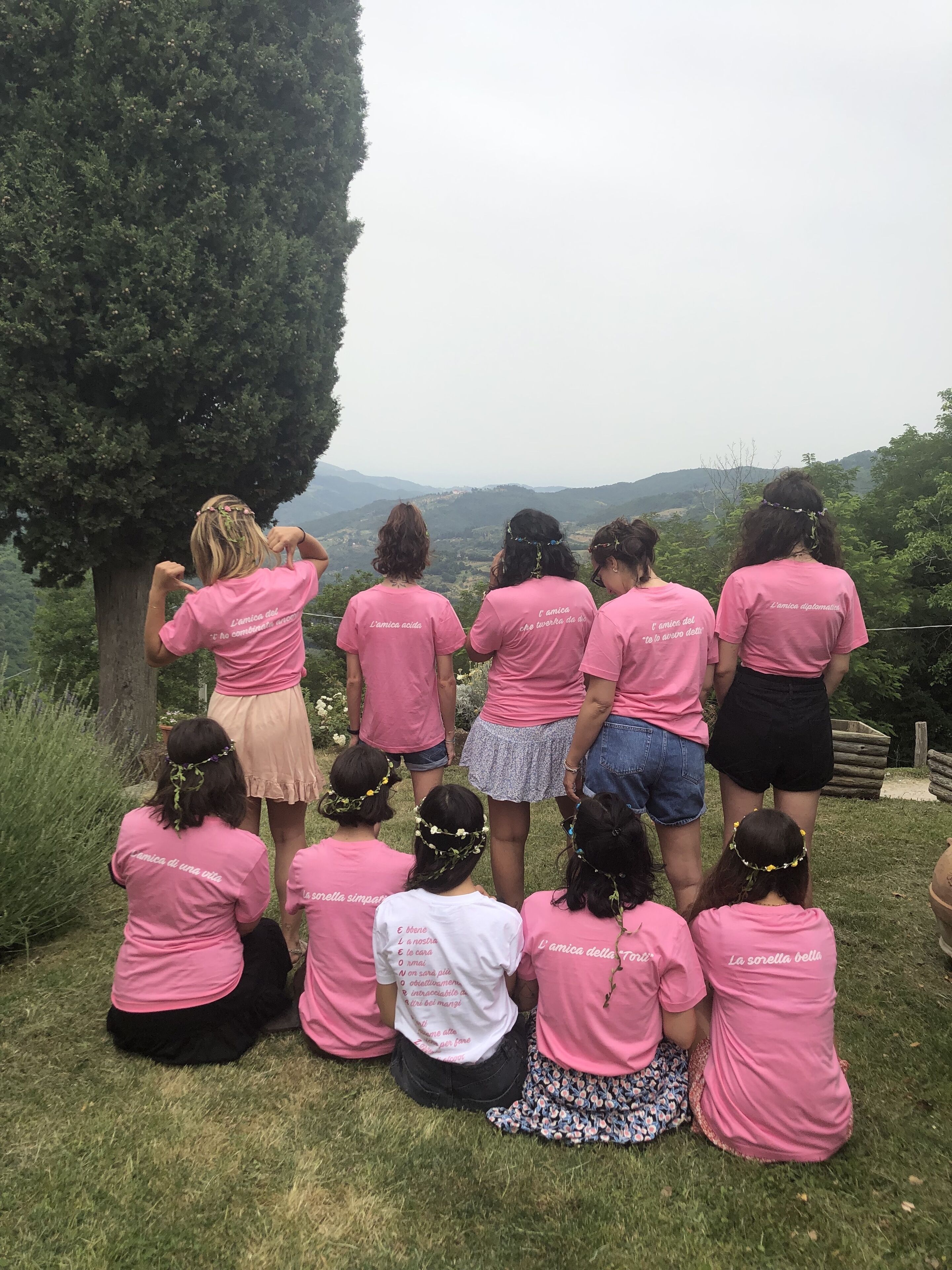 A group of people in pink shirts sitting on grass, overlooking a scenic view of hills and trees.