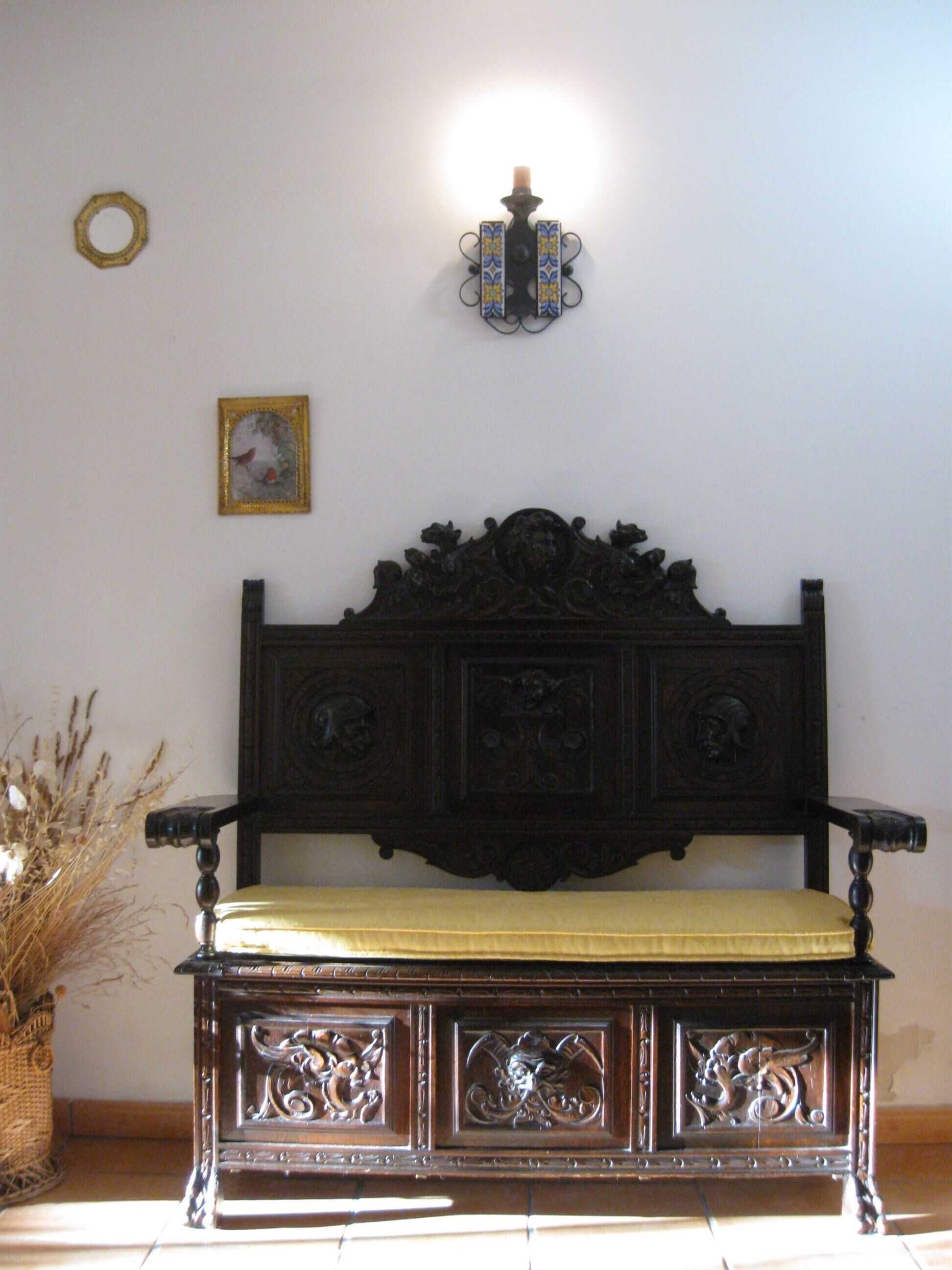 Un banc en bois sombre orné de sculptures complexes, un coussin et un luminaire fixé au mur au-dessus.