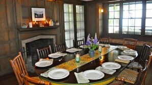 A dining room with a large wooden table set for a meal, flanked by chairs. A fireplace is visible on the left, and a window provides natural light.