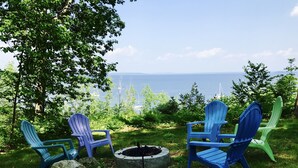 A seating area with Adirondack chairs around a fire pit, overlooking a body of water and trees.