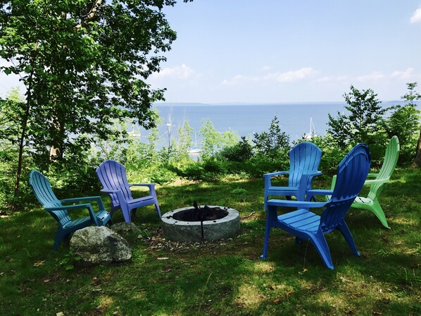 Área de estar con sillas Adirondack alrededor de un brasero, con vista a un cuerpo de agua y árboles.