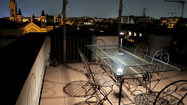 Terraza en la azotea con una mesa de vidrio y sillas de metal, con vistas a un paisaje urbano con edificios iluminados y una torre de iglesia.