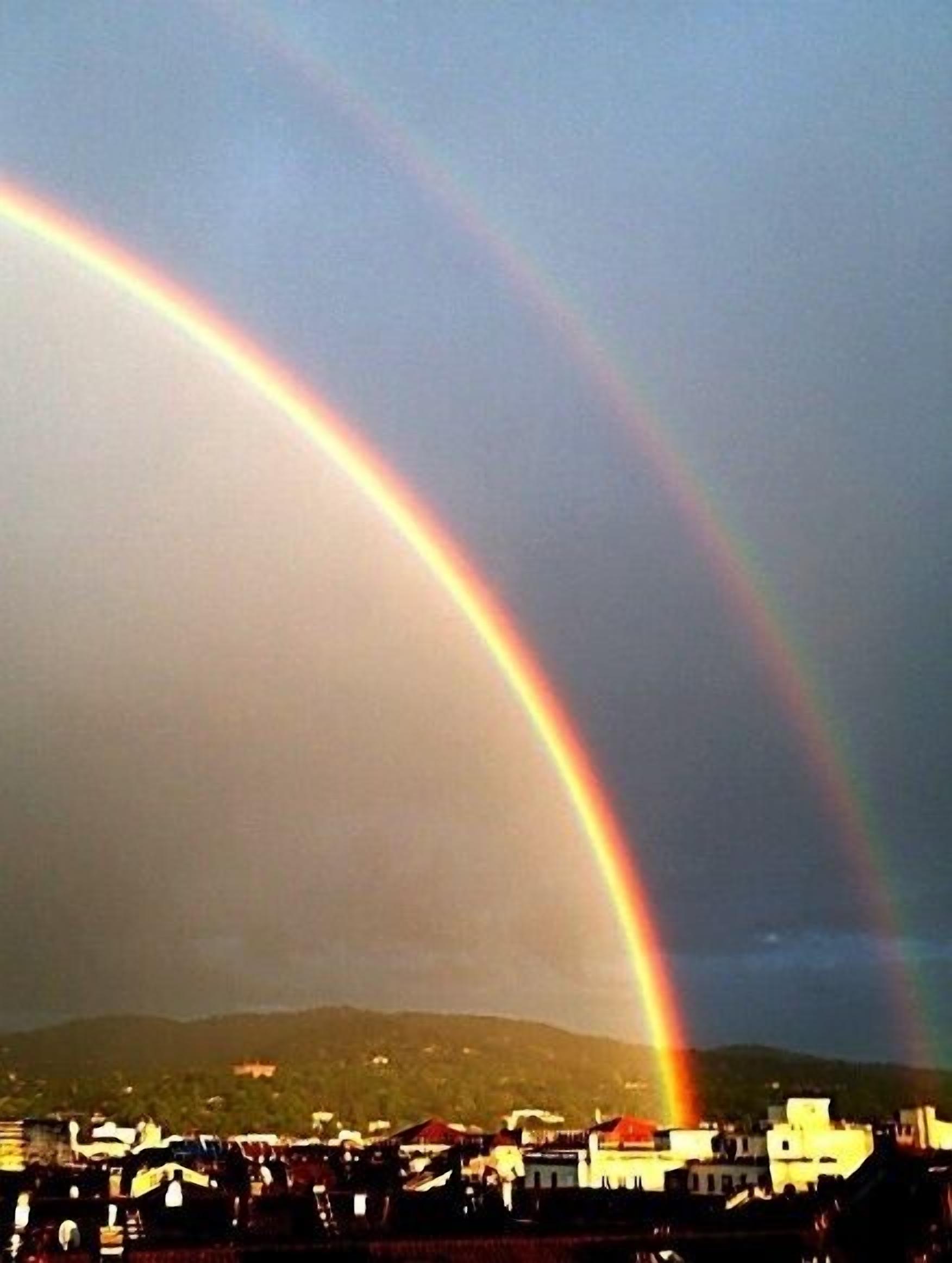 Een dubbele regenboog boven een woongebied met huizen en een heuvelachtig landschap op de achtergrond.