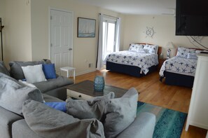 A room with a sofa, two beds with star-patterned bedding, a small table with a vase, a door, and a wall-mounted TV.