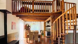 Un amplio salón con techo alto, escalera de madera y una zona de comedor con mobiliario de mimbre.