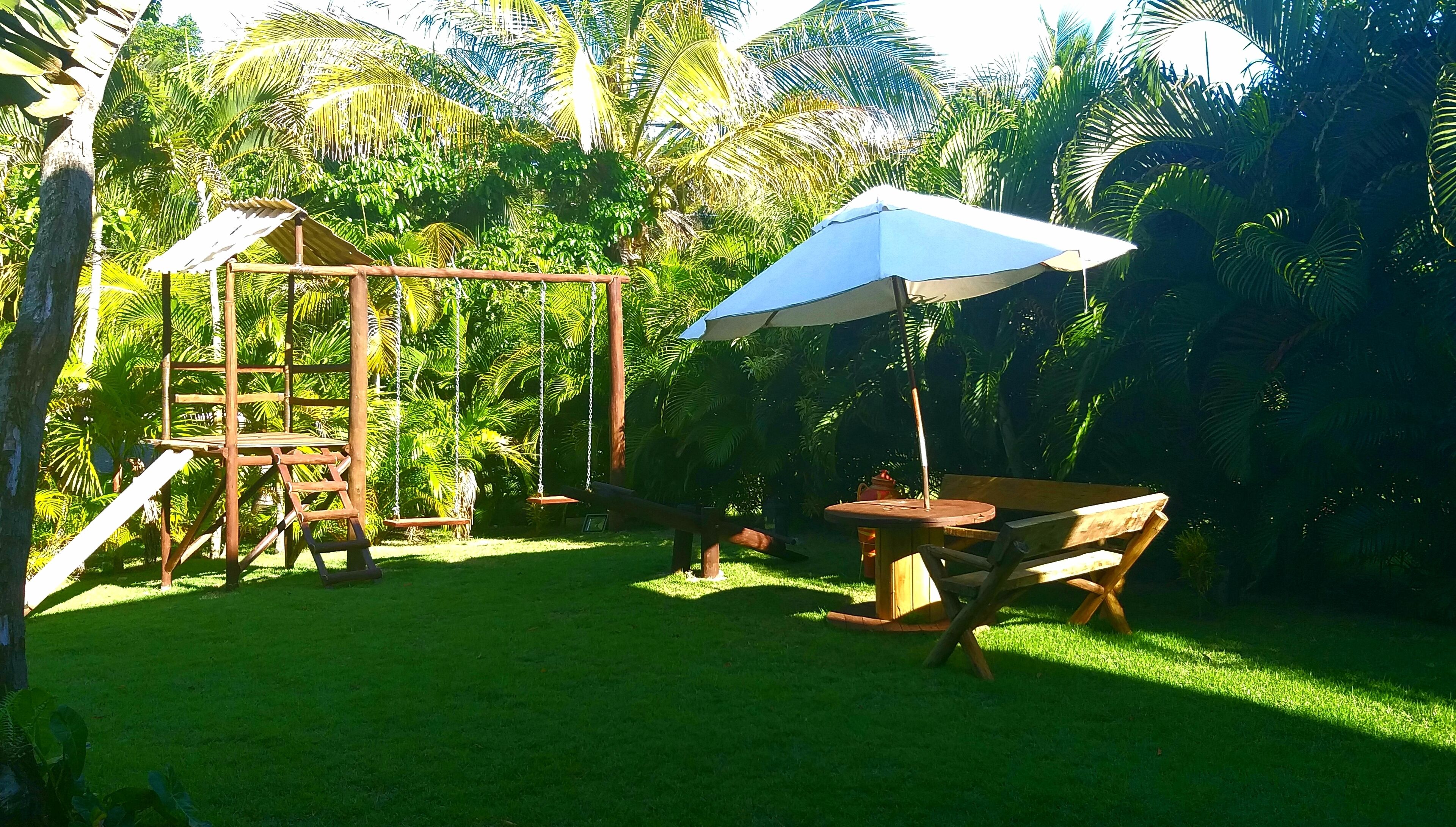 Ein hölzernes Schaukelgerüst, ein Tisch mit Stühlen und ein blauer Sonnenschirm in einem üppigen grünen Garten.