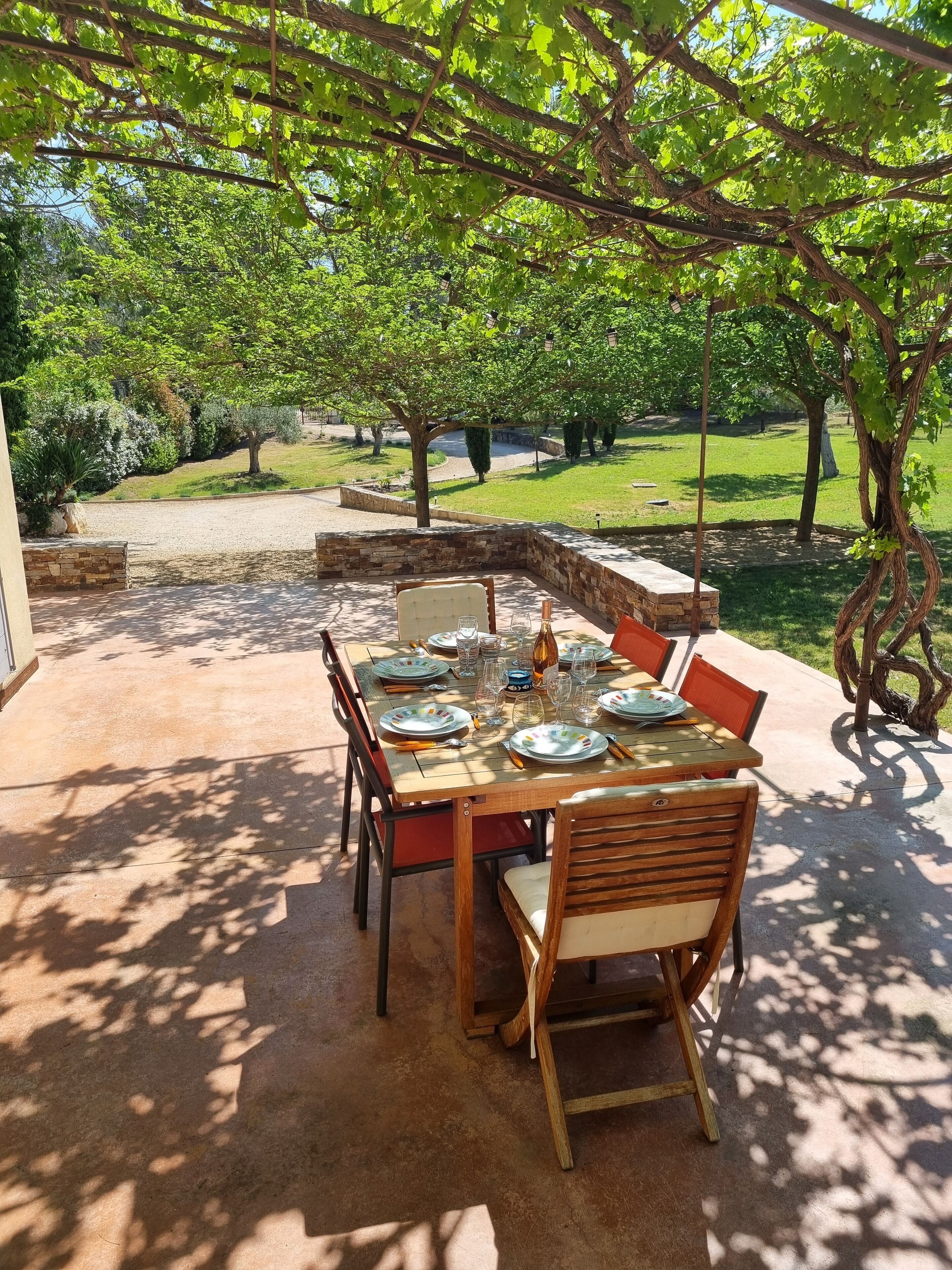 Un'area pranzo all'aperto con un tavolo apparecchiato per quattro, circondato da sedie, sotto una pergola ricoperta di viti.