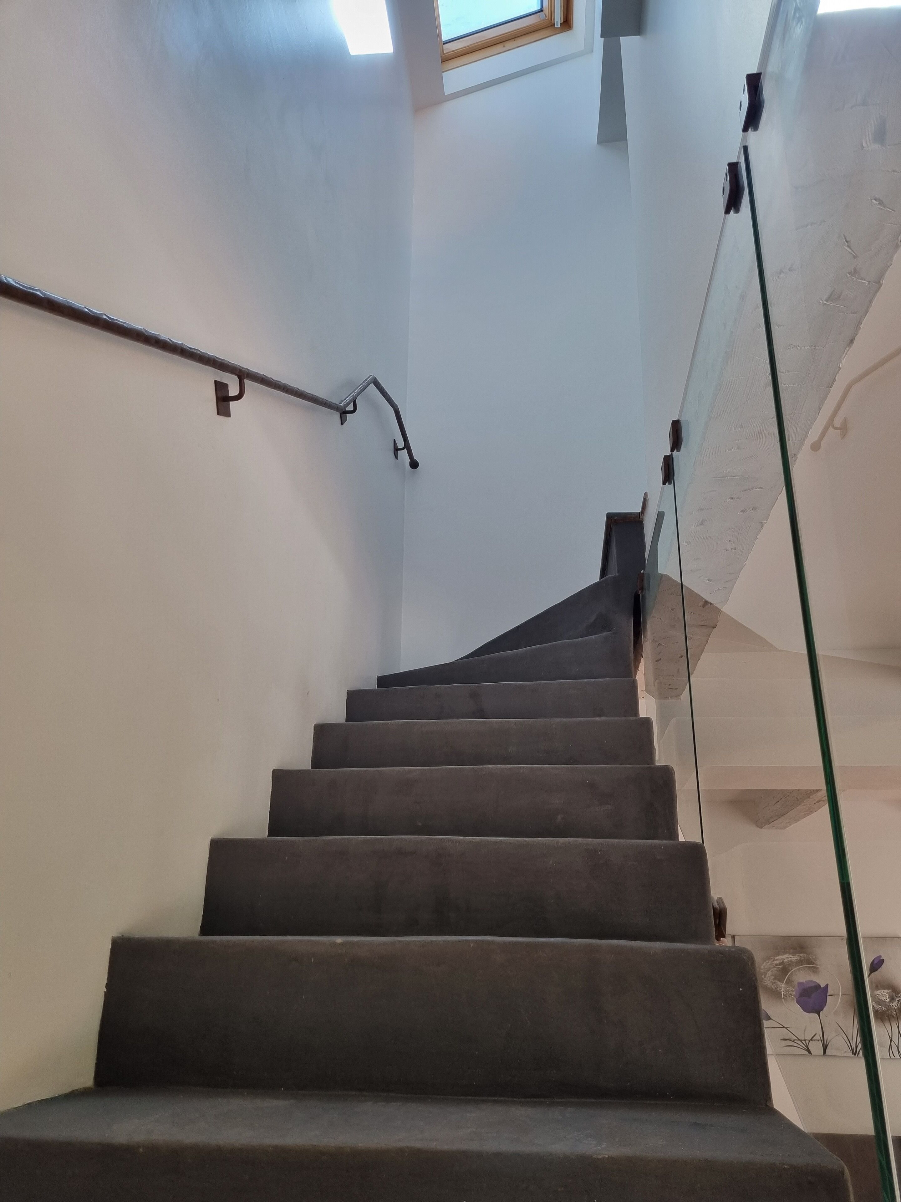 Un escalier doté d’une rambarde en verre et d’une main courante, menant à un palier avec une petite fenêtre.