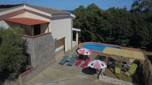 Un área de piscina con sillones de descanso y sombrillas, rodeada por un muro de piedra y un edificio con techo de tejas.