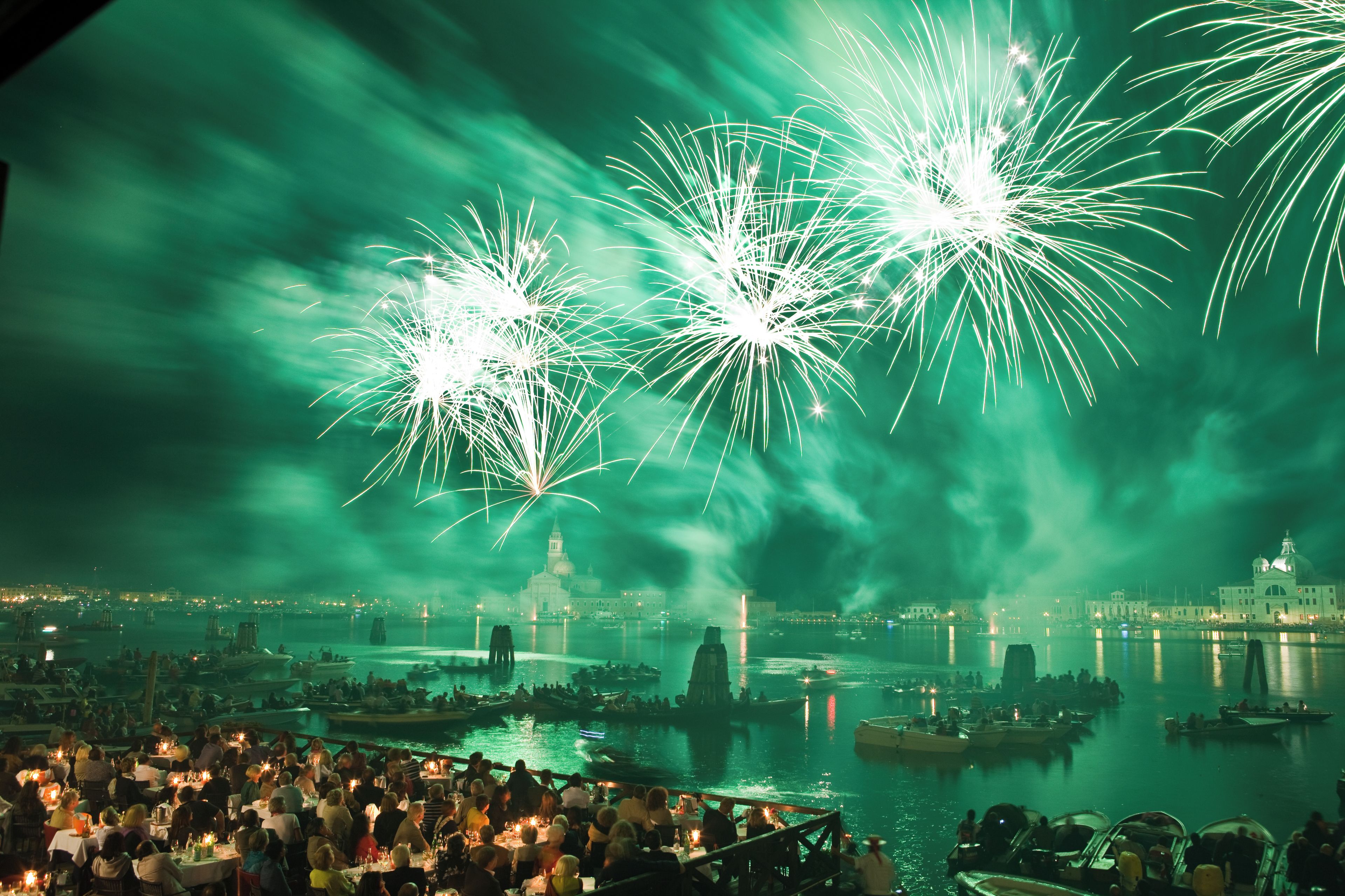 A waterfront cityscape with fireworks, boats, and a crowd of people.
