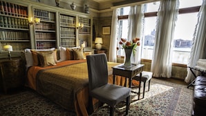 Een kamer met een groot bed, een stoel, een tafel met een bloevas en een boekenplank vol boeken.