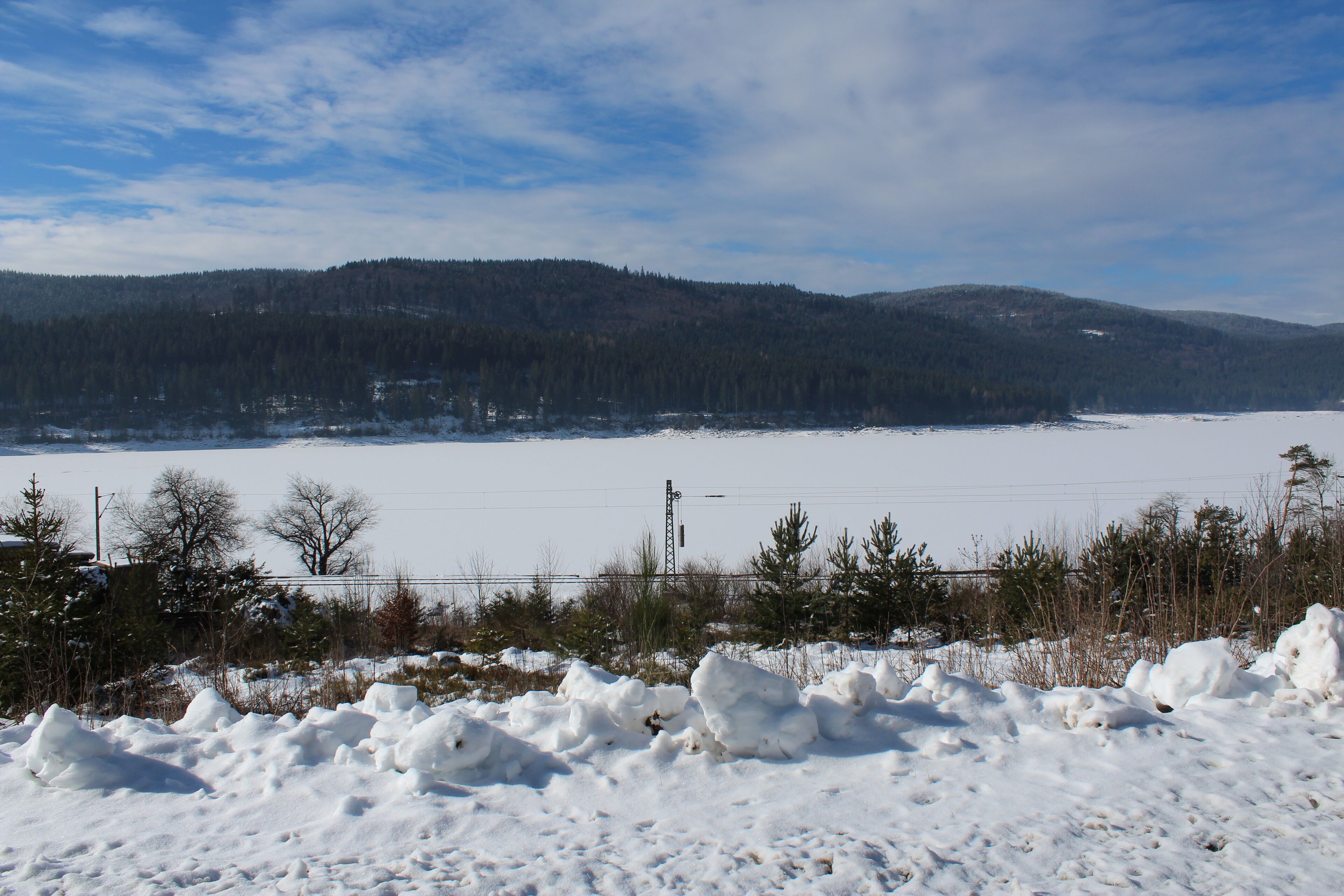 Eine verschneite Landschaft mit einem zugefrorenen See, Bäumen und einer entfernten Hügelkette.