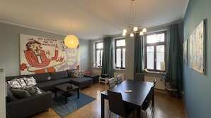 A modern living room with a sofa, coffee table, and dining table. There is a large wall mural, a chandelier, and large windows with curtains. - Family-friendly 3-room apartment in the middle of hip Pieschen (Dresden)