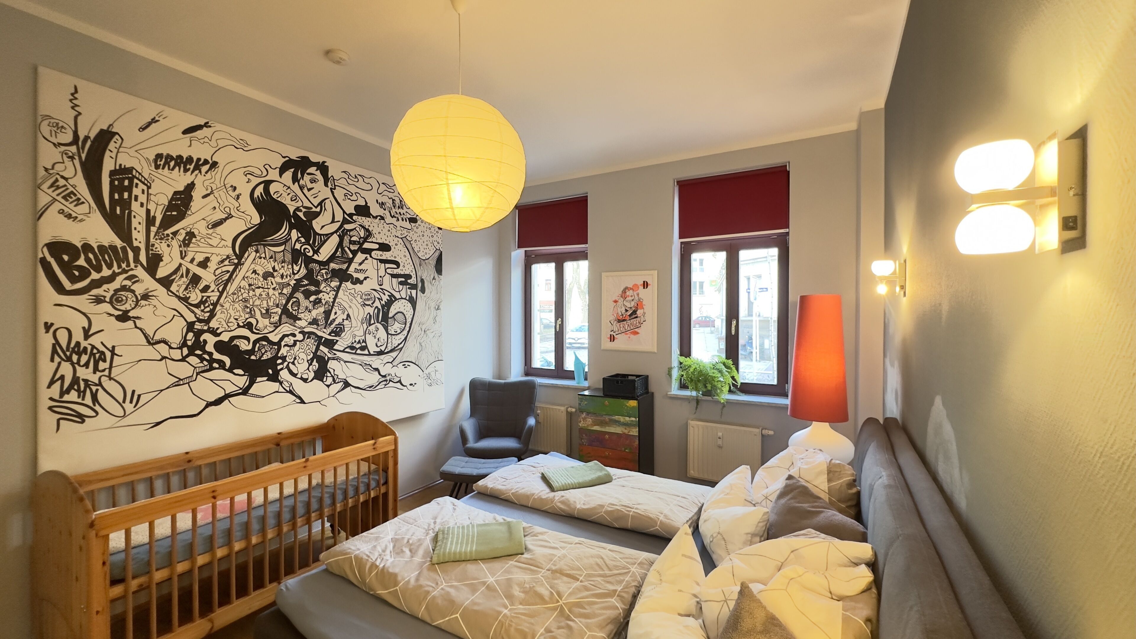 Ein Schlafzimmer mit zwei Betten, einem Holz-Babybett, einem großen Wandbild, einem Sessel, einem Heizkörper und einem Fenster mit Blick auf die Straße.