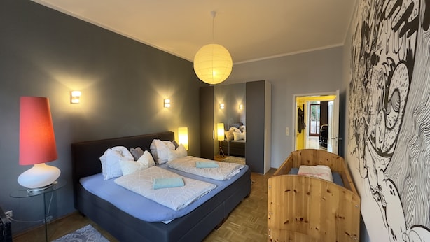 A bedroom with a bed, bedside lamps, a wooden chair, and a wall with a large black and white mural. - Family-friendly 3-room apartment in the middle of hip Pieschen (Dresden)
