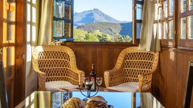 A room with a wooden ceiling, two wicker chairs, a table with a tray of bread, and a view of mountains through a window.