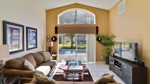 A living room with a sofa, coffee table, TV, and a large window overlooking a pool. - Popular 4 bedroom pool home is a must for family fun, located in Emerald Island (Kissimmee)