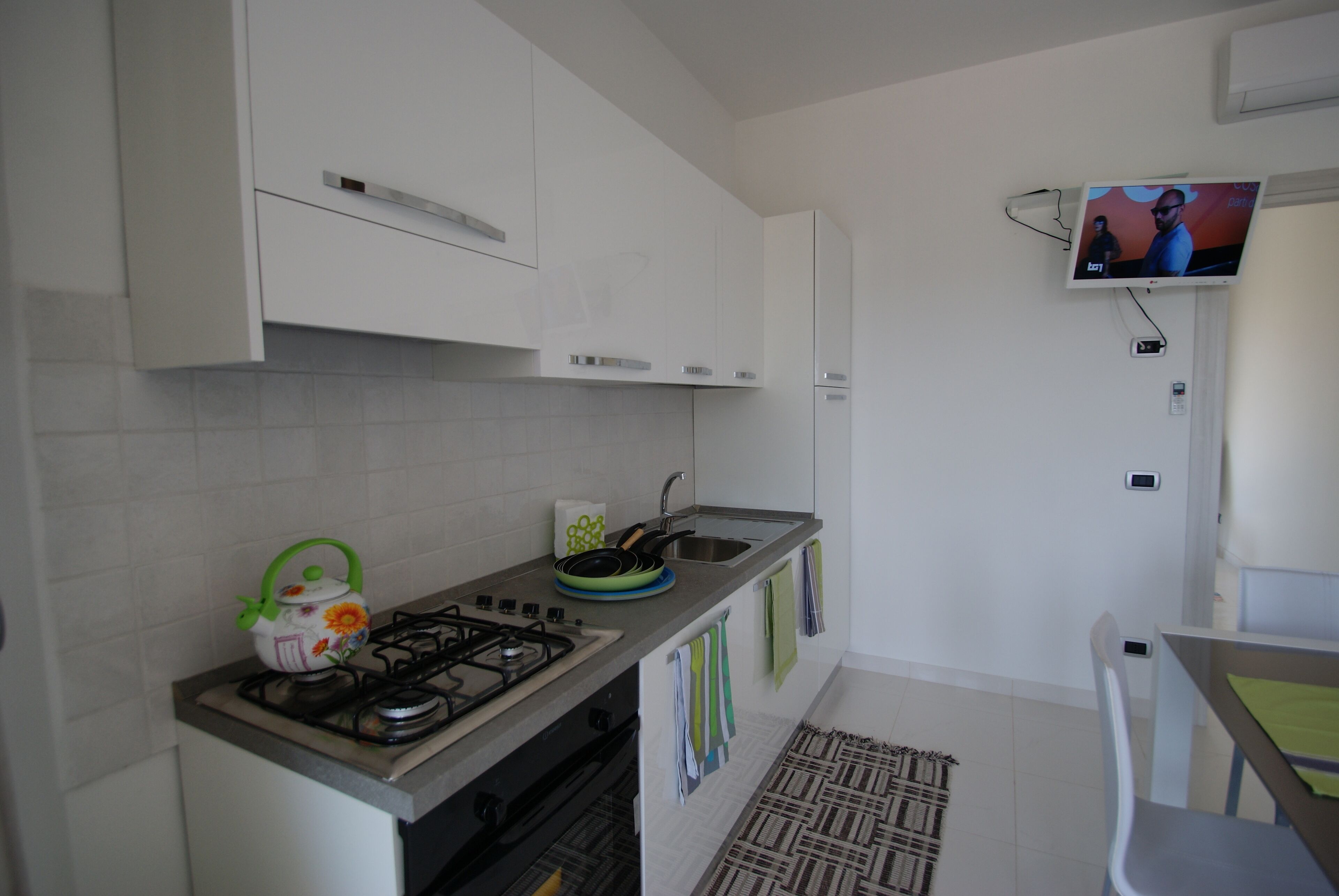 A modern kitchen with white cabinets, a gas stove, a kettle, and a flat-screen TV mounted on the wall.