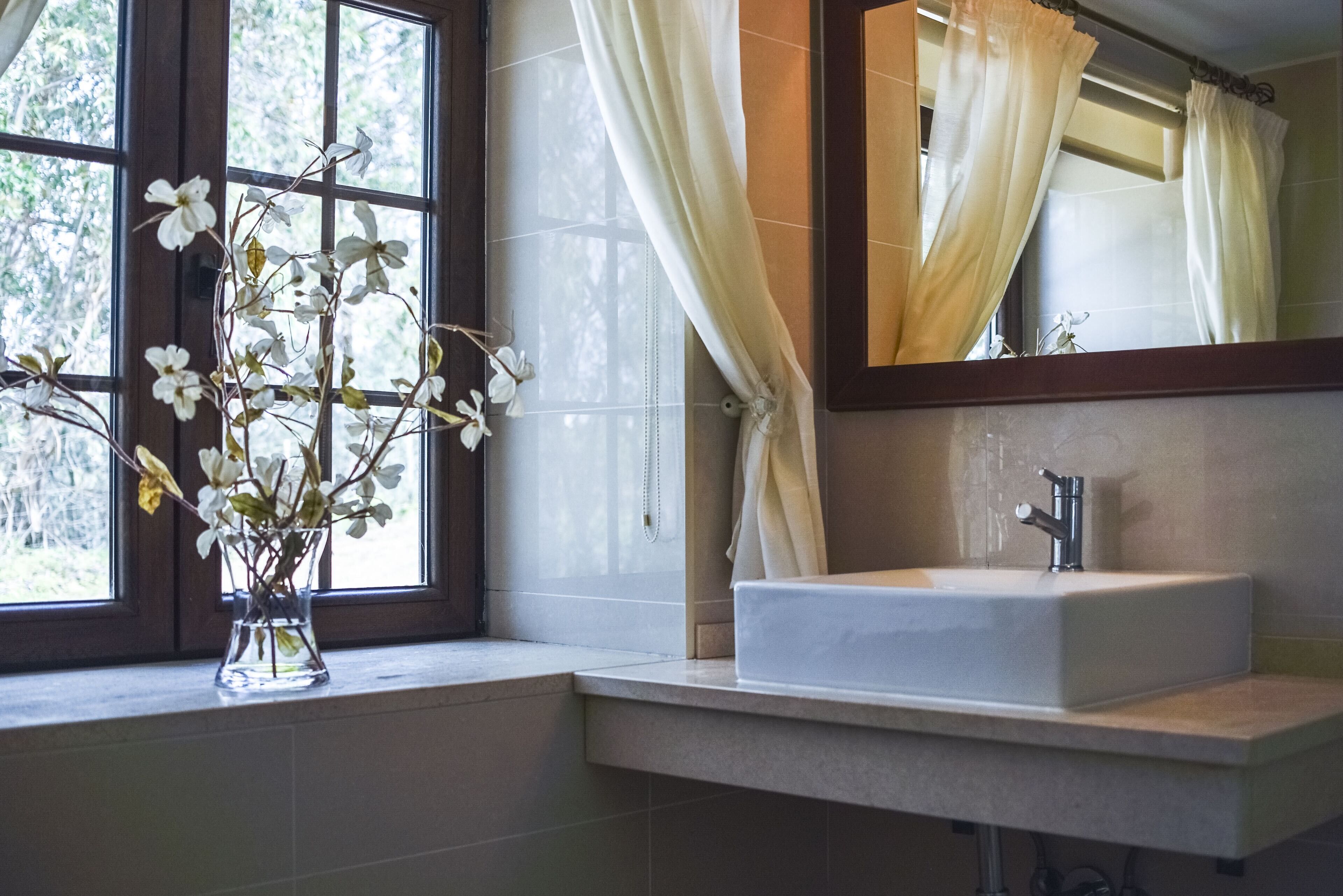 A bathroom with a window, a vase with flowers, a sink, and a mirror.