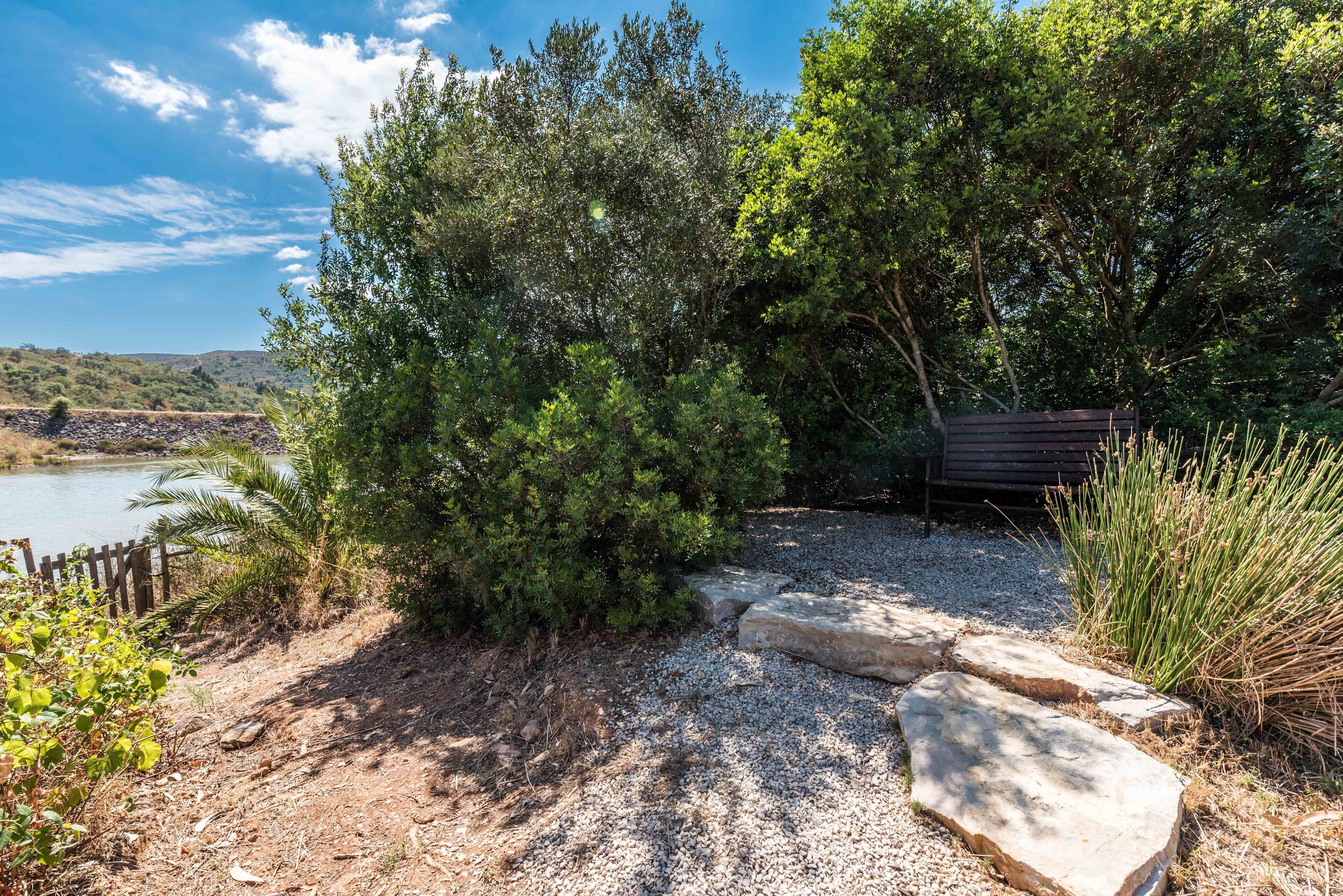Un chemin de gravier, ponctué de pierres plates, traverse un jardin où la végétation est luxuriante, avec de nombreux arbustes et arbres.