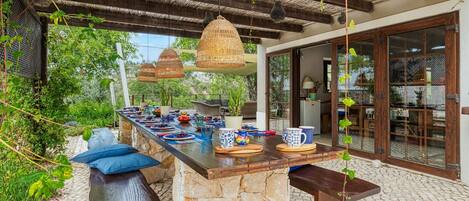 An outdoor dining area with a stone island, wooden benches, and hanging wicker lamps.