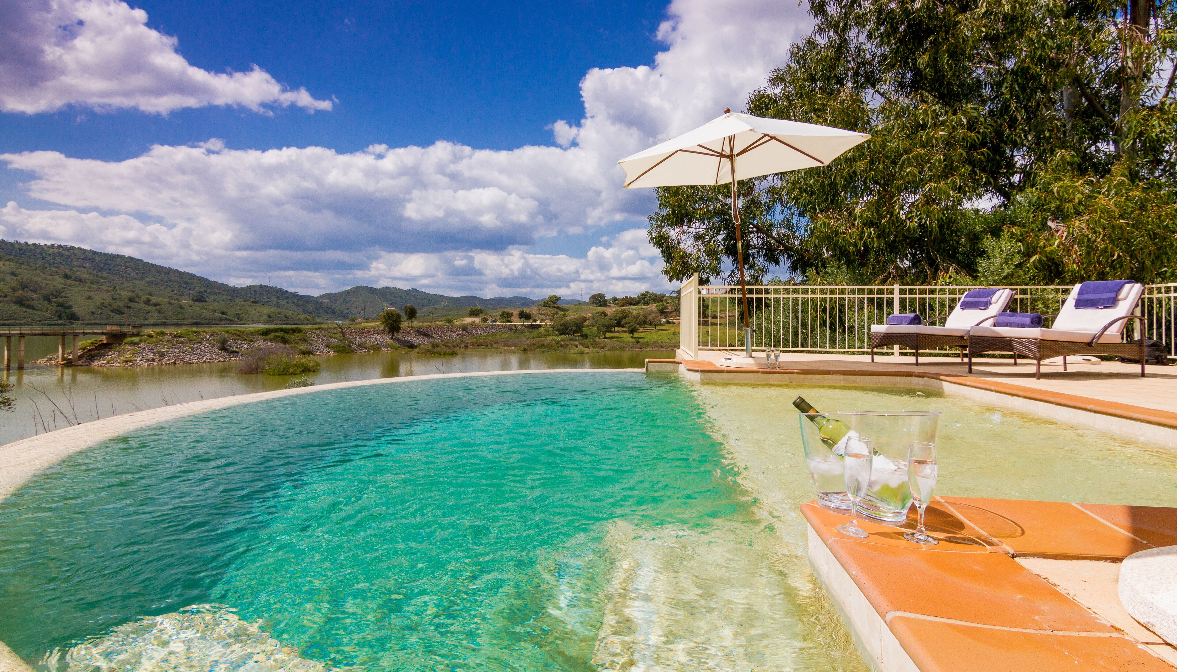 A clear pool with a white umbrella, lounge chairs, and a view of hills in the background.