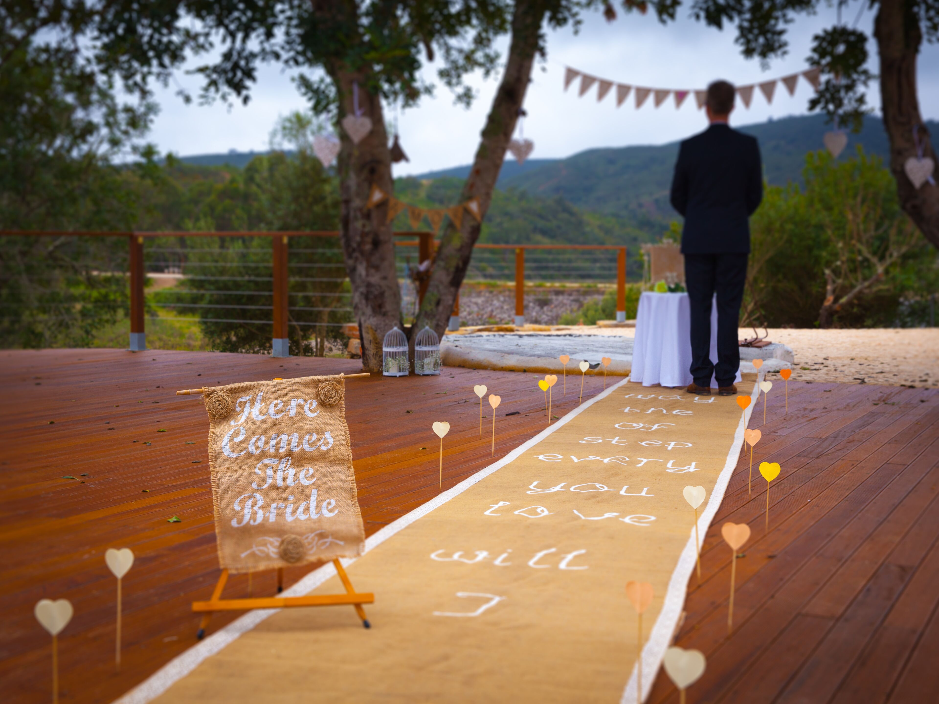 Een houten terras met een witte tafel en een bord waarop staat 'Here Comes the Bride'.
