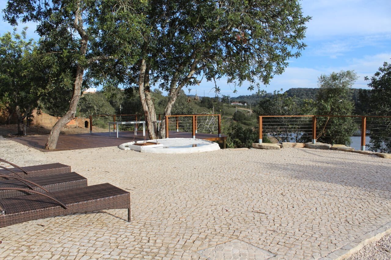 A paved outdoor area with a circular water feature, wicker lounge chairs, and a view of a body of water.