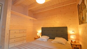 Un dormitorio de cabaña de madera con una cama, dos mesitas de noche con lámparas, un aparador y un cuadro en la pared.
