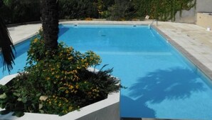 A rectangular swimming pool with clear blue water, surrounded by a white pool deck and bordered by a low hedge.
