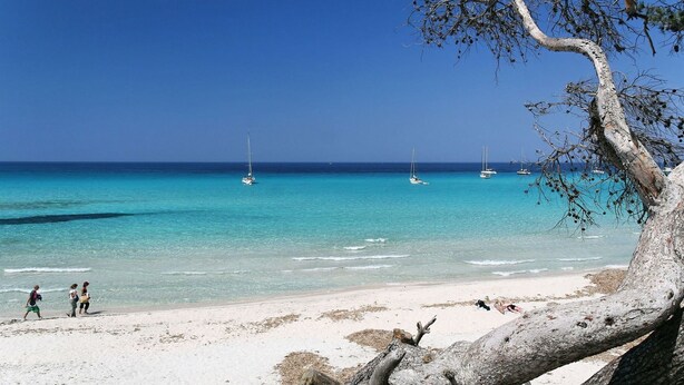 A beach with clear turquoise water, a few sailboats, and people walking on the shore.