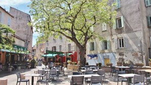 An outdoor café area with tables and chairs, surrounded by buildings with shutters and signage.