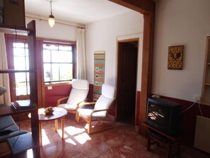 Ein Wohnzimmer mit Holzboden, zwei weiße Sessel, ein kleiner runder Tisch, einem Fernseher und einem Fenster mit Vorhängen.