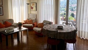 A living room with a sofa, armchairs, a coffee table, and a dining table. There is a window with curtains, a radiator, and a painting on the wall. - Apartment - Bellagio House of the Sun (Bellagio)