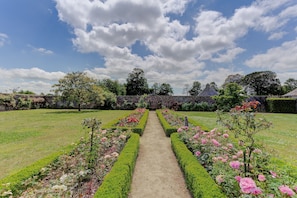 Un chemin de jardin bordé de haies soigneusement taillées et de plantes en fleurs, qui mène à un mur de pierre et à des arbres en arrière-plan.