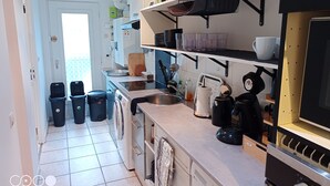 A modern kitchen with a dishwasher, washing machine, stove, and various kitchen utensils. There are two trash bins and a window in the background.