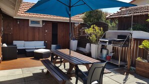 En udendørs terrasse med et træbord og bænke, en blå parasol og en grill.