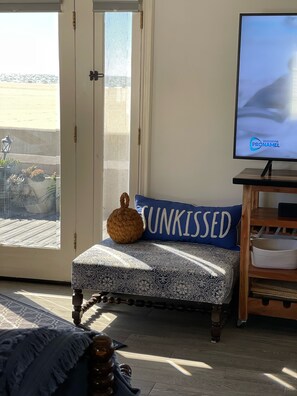 A room with a patterned ottoman, a television displaying a logo, and a view through a door.