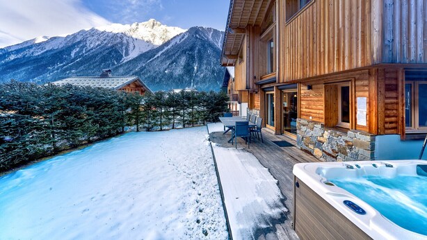 Een houten lodge met een hottub, een terras met een tafel en stoelen, en uitzicht op besneeuwde bergen.