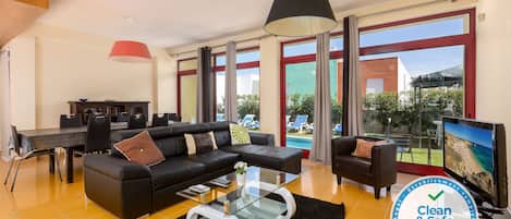 A modern living room with a black sofa, a glass coffee table, and a TV. There is a dining table with chairs and a red pendant light. Large windows offer a view of the outdoors.